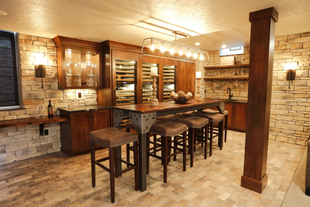 Rustic modern home bar with stone walls, wooden cabinetry, large wooden bar table with cushioned stools, glass-front cabinets, wine refrigerator, and warm ambient lighting.