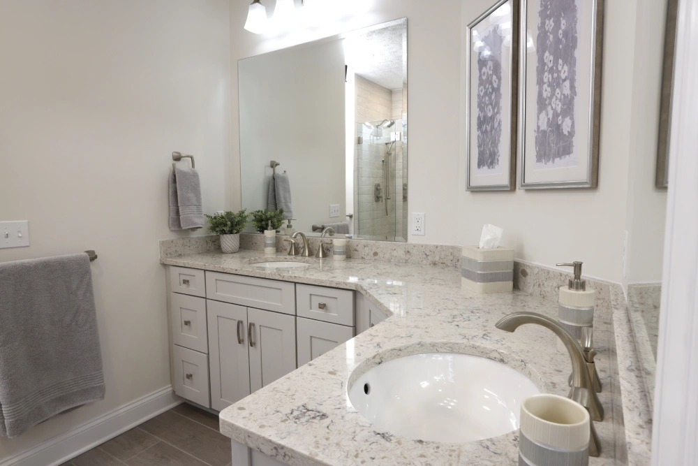 Modern bathroom with granite double sink countertop, brushed nickel faucets, white cabinets, large mirror, decorative items, floral wall art, and light gray tiled floor.
