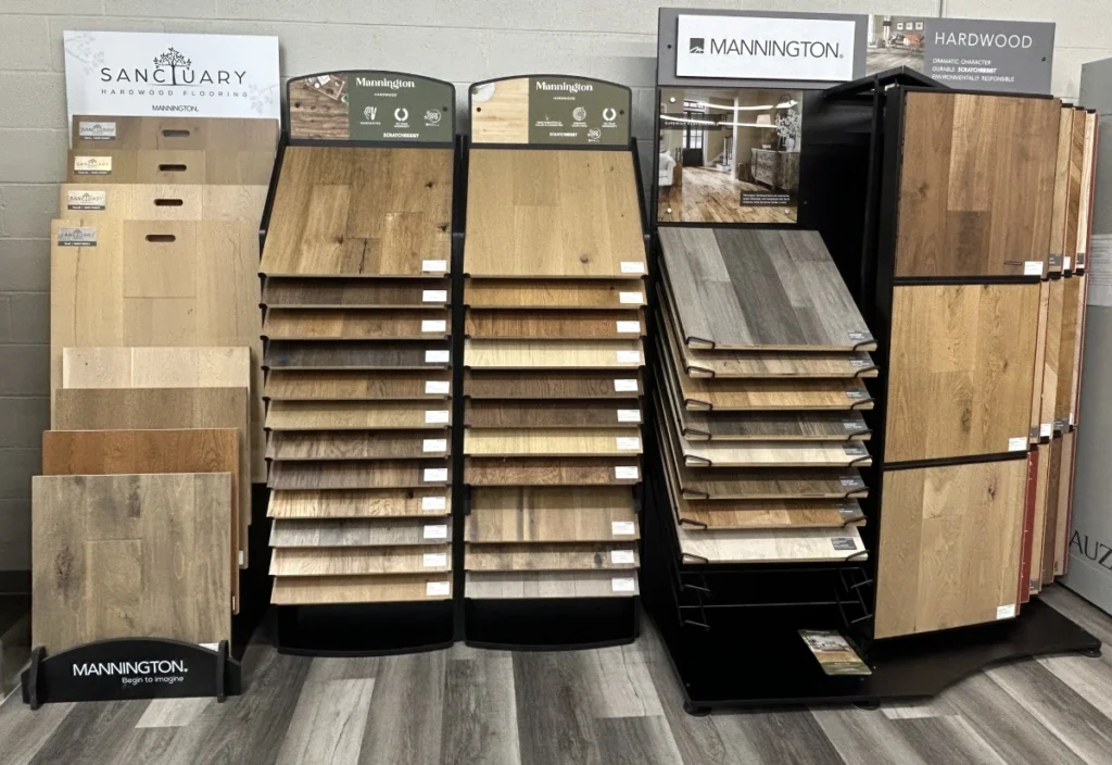 Showroom display of hardwood flooring samples with multiple racks of wood planks in light, dark, and gray tones, featuring Sanctuary and Mannington branded sections.