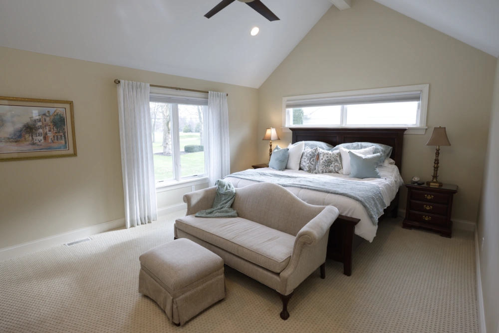 Bedroom with vaulted ceiling, dark wooden headboard, white bedding, blue and white pillows, beige loveseat with ottoman, nightstands with lamps, large window with curtains, and light-colored carpet.
