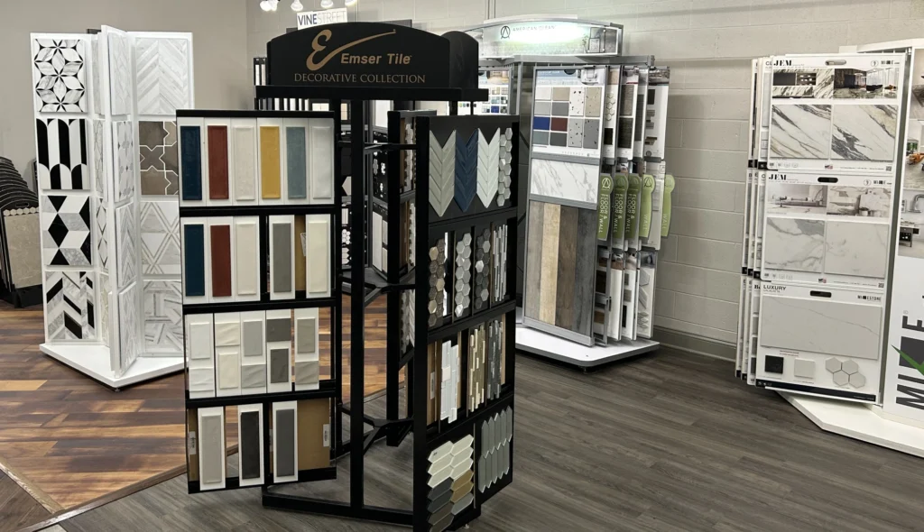 Showroom display of decorative tile samples on Emser Tile rotating stand, surrounded by marble, wood-look, and geometric flooring designs.