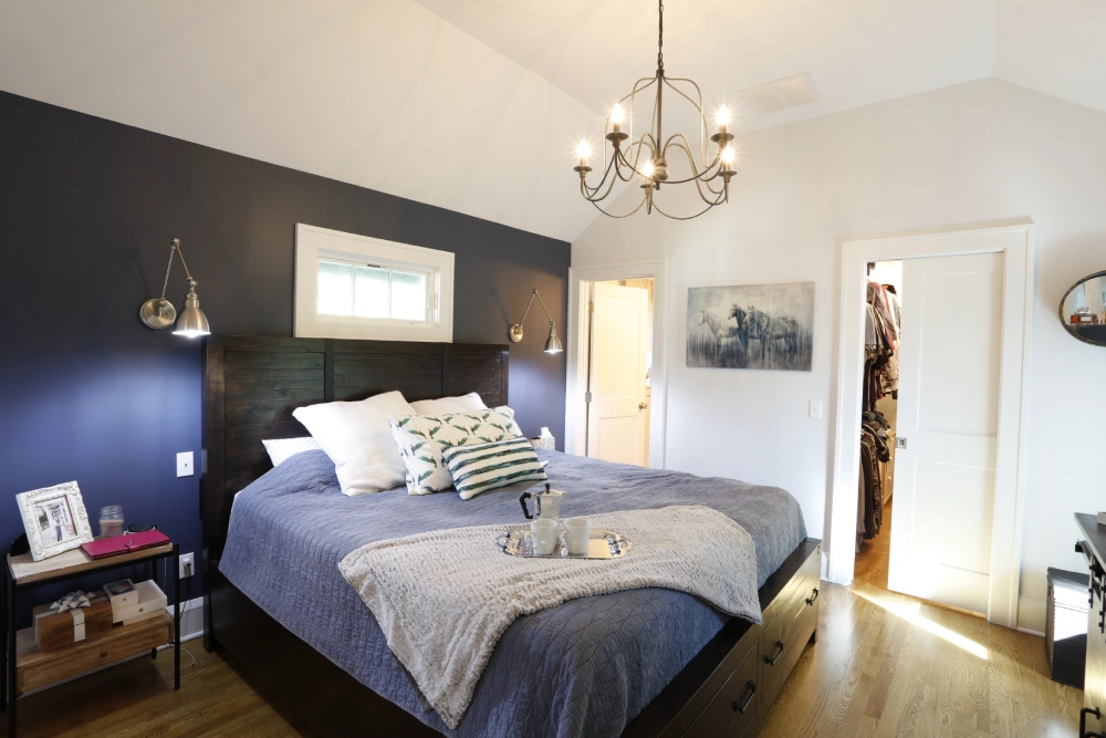Modern bedroom with dark wooden bed frame, blue quilted comforter, pillows, gray throw blanket, pendant lights, chandelier, bedside table, horse painting, and wooden flooring.