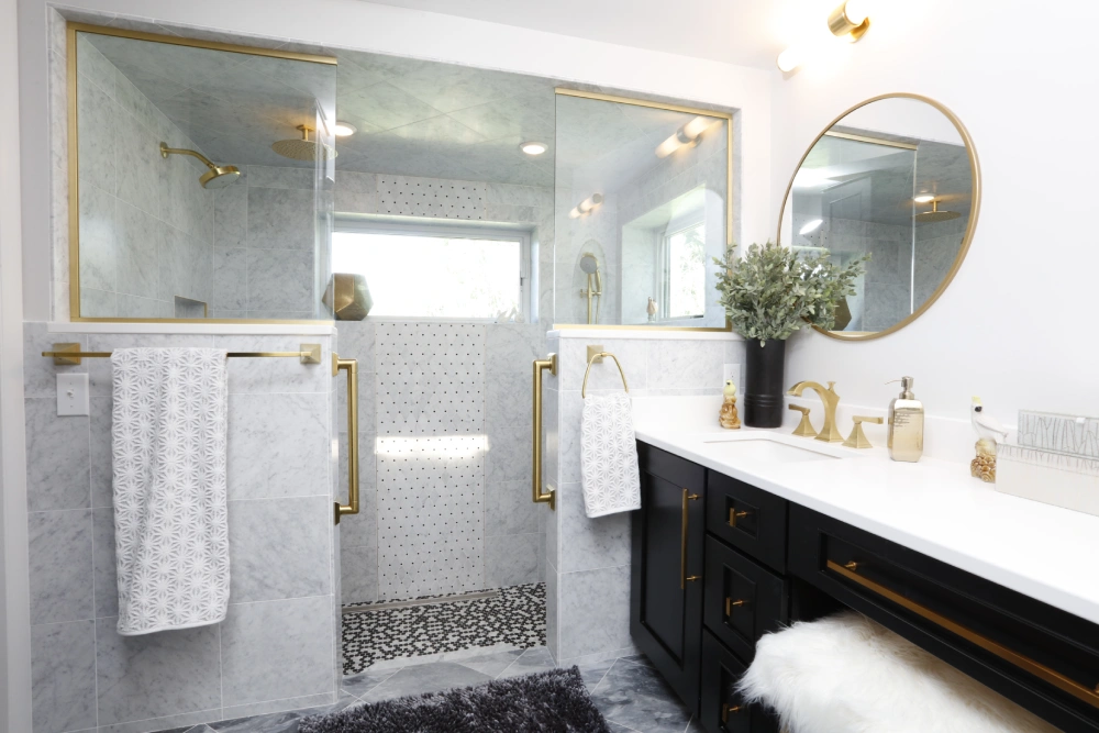 Luxurious modern bathroom with glass-enclosed shower, gold trim and fixtures, marble tiles, patterned mosaic floor, white vanity with black cabinetry, round gold-framed mirror, and decorative countertop items.