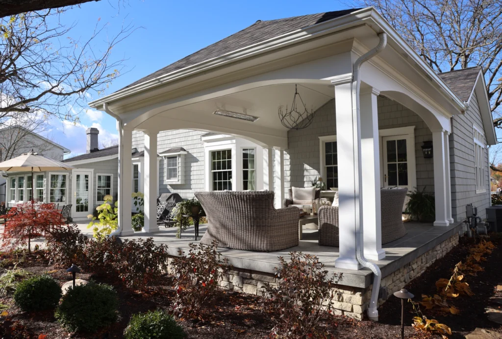 Residential backyard patio with covered seating area, white columns, wicker chairs, chandelier light, landscaped garden, and gray shingle roof.